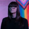 person wearing glasses and black jacket standing before pride flag