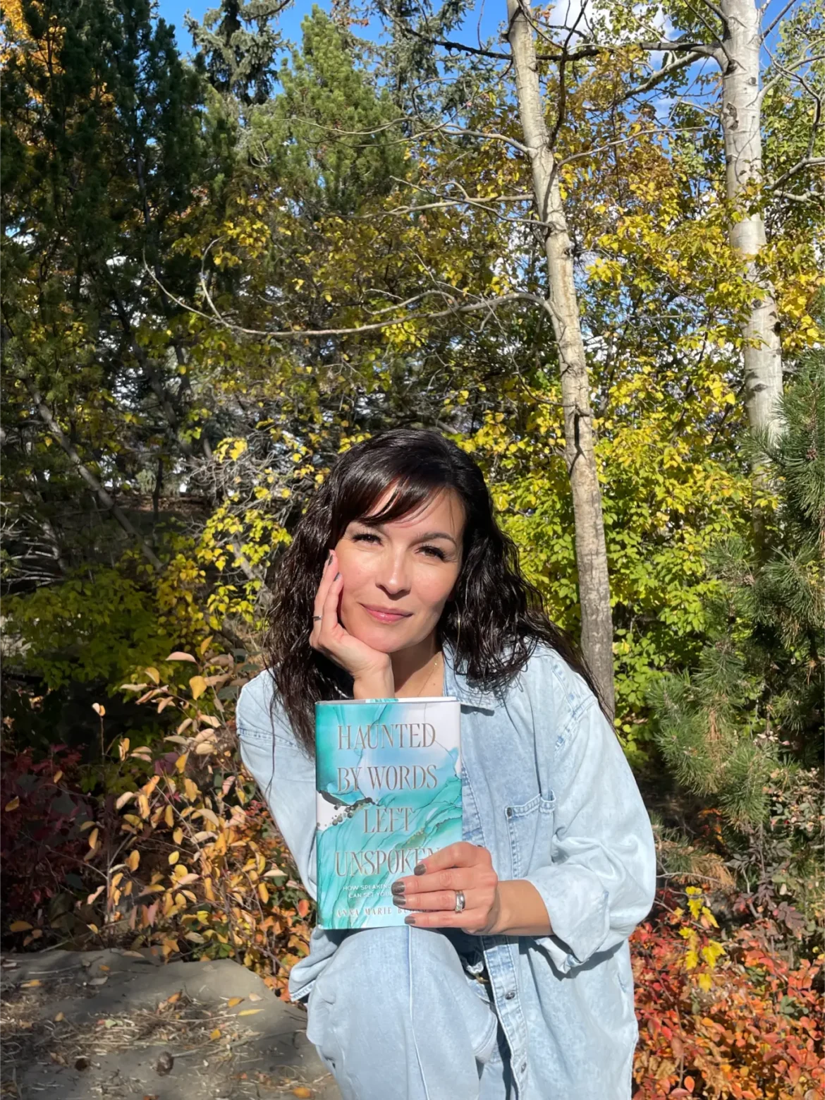 Woman holds "Haunted by Words Left Unspoken" book outdoors amidst fall foliage.