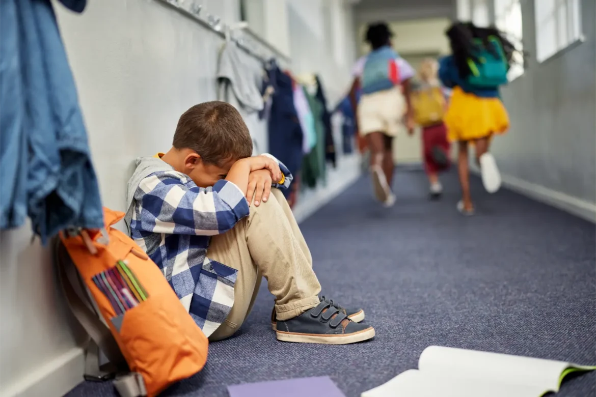Sad boy sitting alone in school hallway, other children running past.