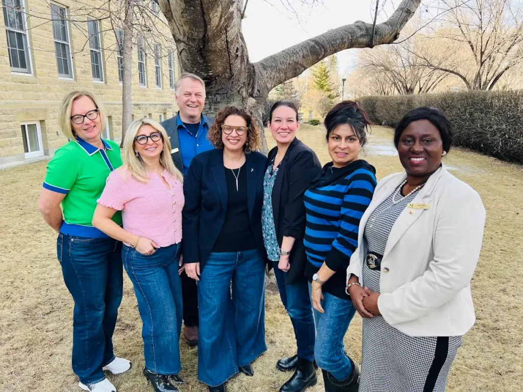 Diverse group of seven people posing for a photo outdoors by a tree and building.