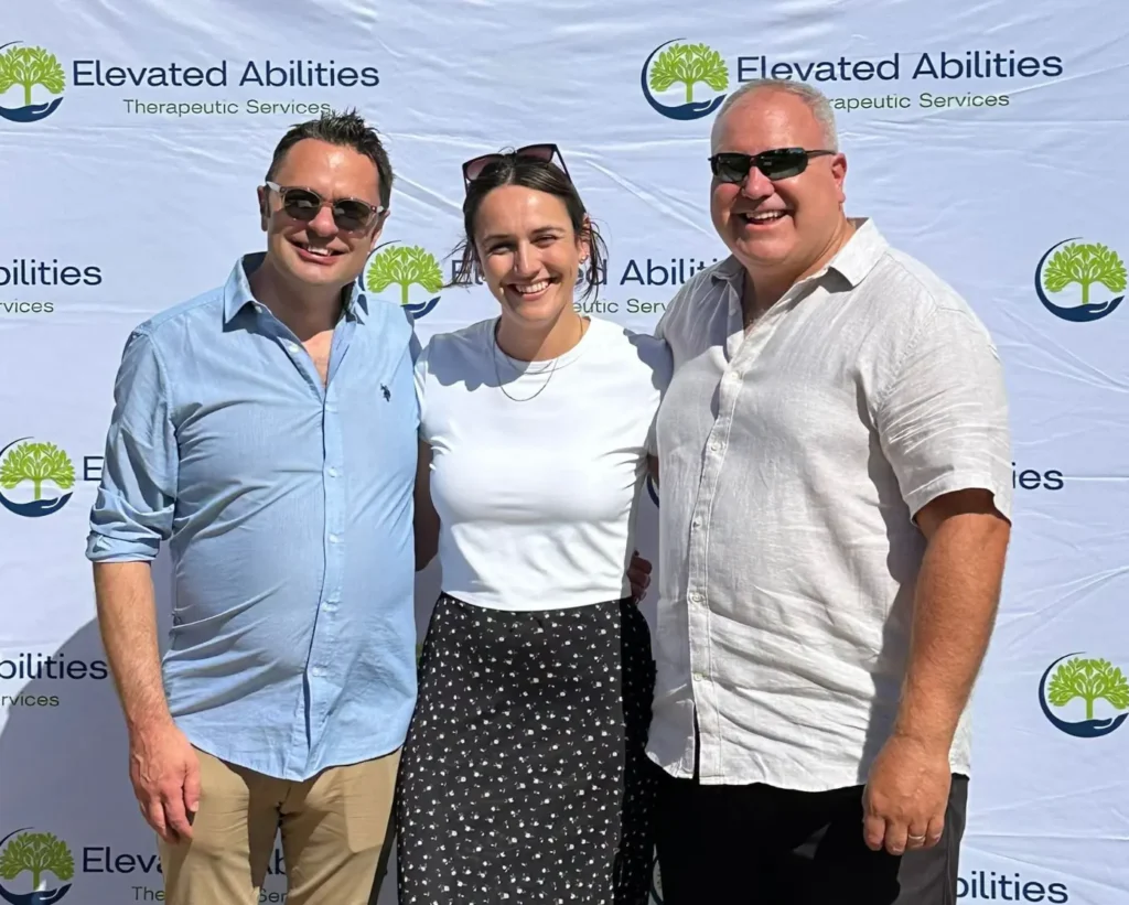 Three smiling people pose before a banner for Elevated Abilities Therapeutic Services.