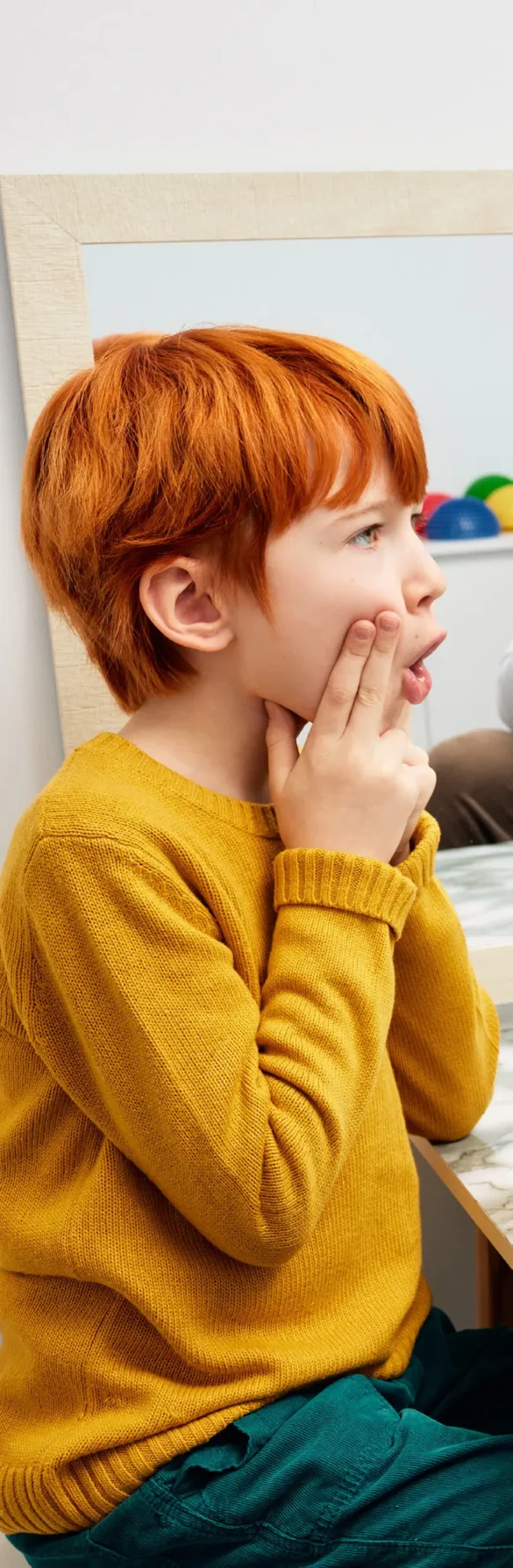 Ginger haired boy practising speech sounds at a mirror with therapist.