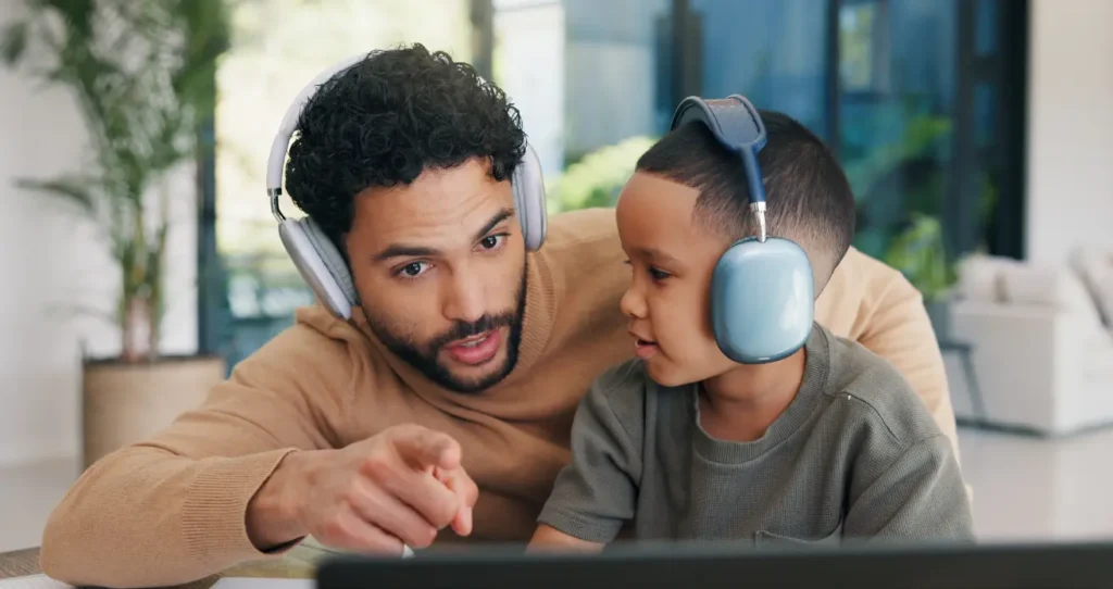 Man wearing headphones points at computer screen assisting a boy wearing headphones