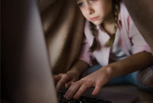 Girl in pink shirt with braided hair typing on a laptop under a blanket fort.