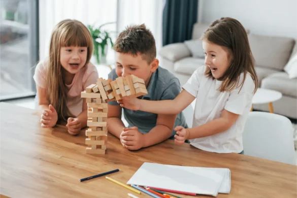 Three children playing Jenga game on wooden table, building a tower of blocks.
