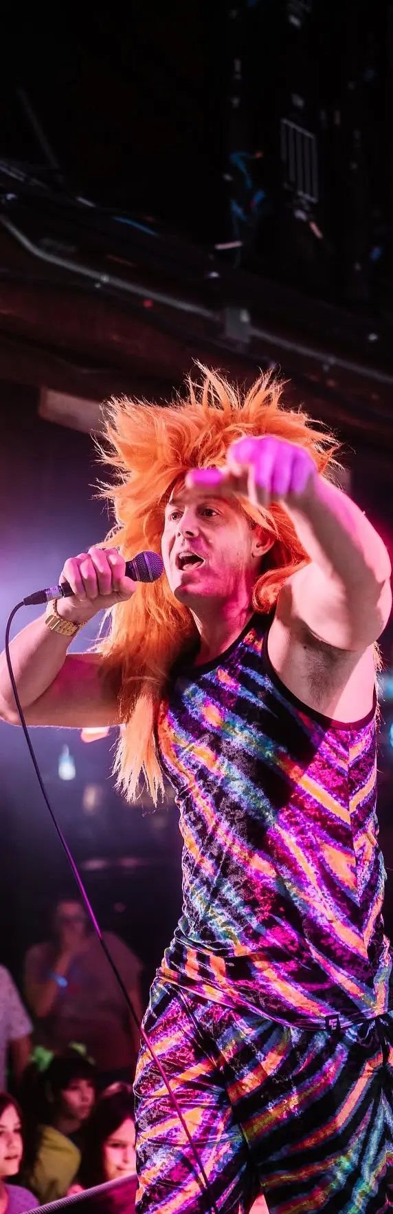 Entertainer in orange wig and colourful outfit performs on stage with a microphone.