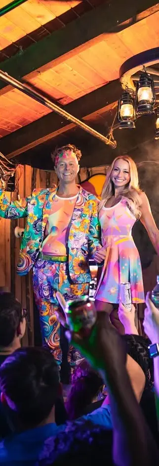 Couple in vibrant psychedelic outfits celebrating with drinks at a concert.