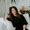 Smiling woman with curly hair in a black shirt sitting on a white couch with plants.