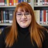 Woman with red hair and glasses smiles in front of bookshelves.