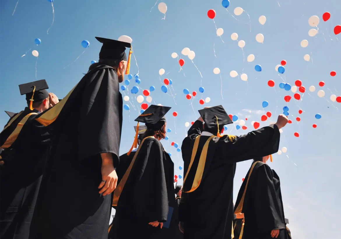 Graduates in black gowns and caps release red, white, and blue balloons into the sky.