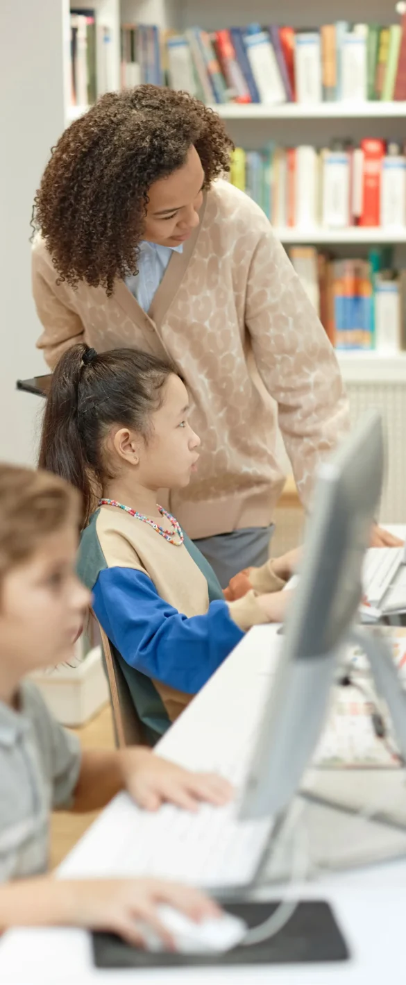 Teacher helping a young student with computer work in a classroom library.