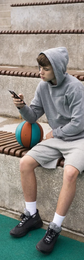 Young man in grey hoodie and shorts with basketball and phone on bleachers