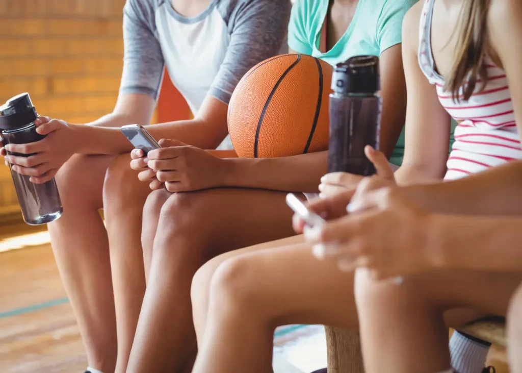 Teenage girls in sports gear with basketball and phones on a bench.