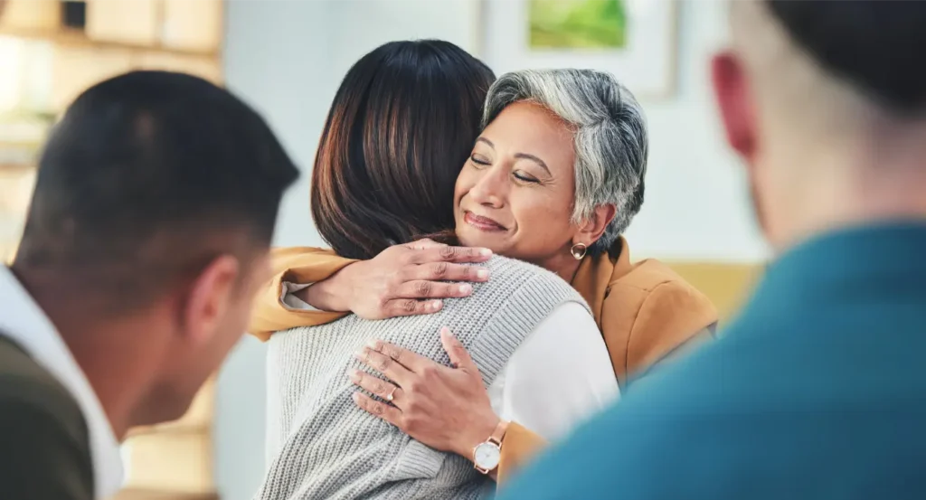 Elderly woman with grey hair joyfully hugs younger woman, two men in foreground blurred.