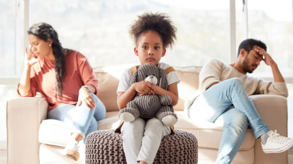 Child clutches stuffed animal between parents looking distressed on sofa