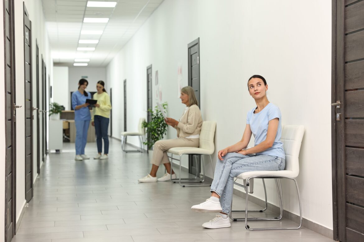 Woman waits in clinic hallway, with nurse and patient in background.