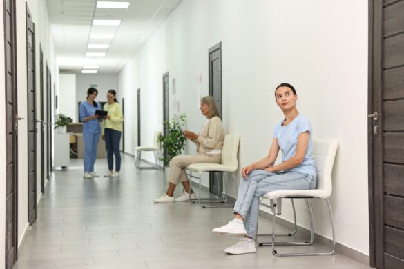 Woman waits in clinic hallway, with nurse and patient in background.
